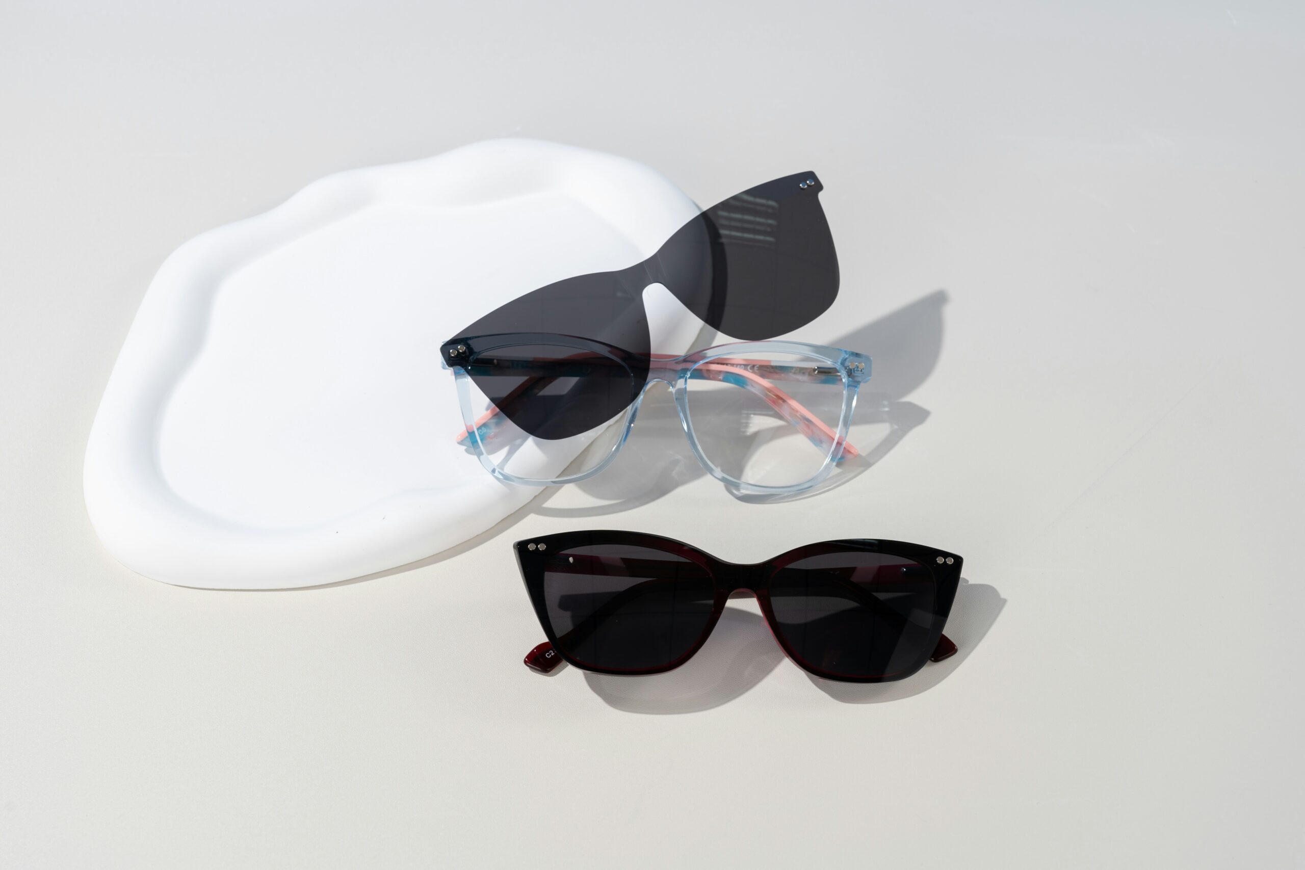 Three pairs of sunglasses on a light surface, including black cat-eye frames, translucent blue frames with pink arms, and oversized black frames.